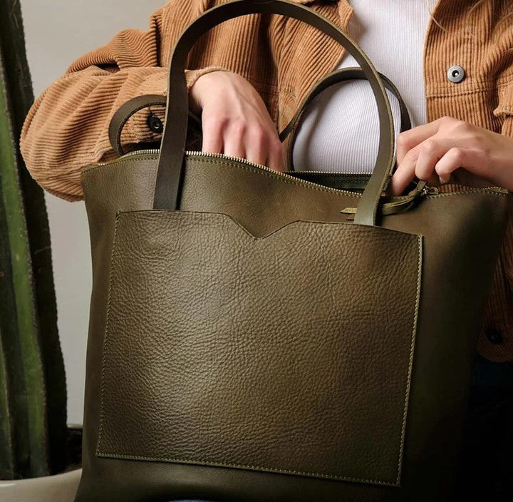 A woman in a tan jacket and white top carries an olive green leather tote bag, showcasing its spacious design and everyday versatility