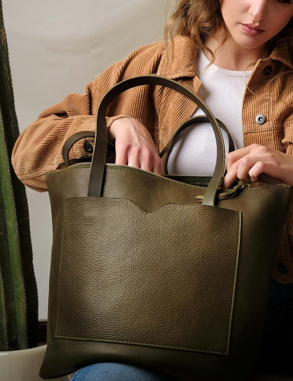 A woman in a tan jacket and white top carries an olive green leather tote bag, showcasing its spacious design and everyday versatility