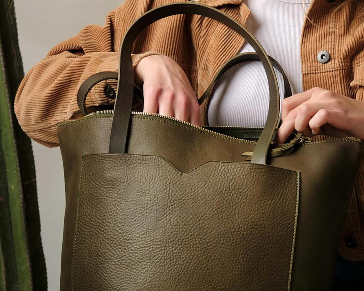 A woman in a tan jacket and white top carries an olive green leather tote bag, showcasing its spacious design and everyday versatility