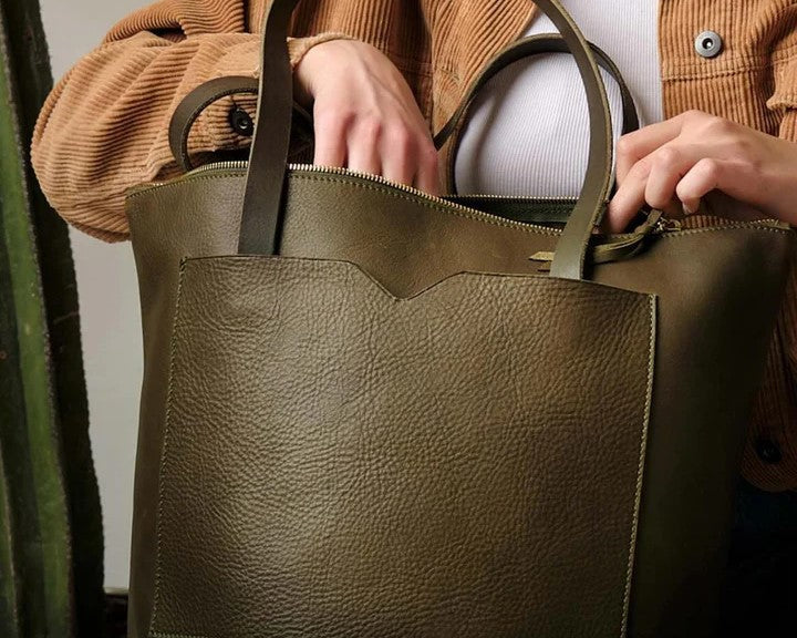 A woman in a tan jacket and white top carries an olive green leather tote bag, showcasing its spacious design and everyday versatility