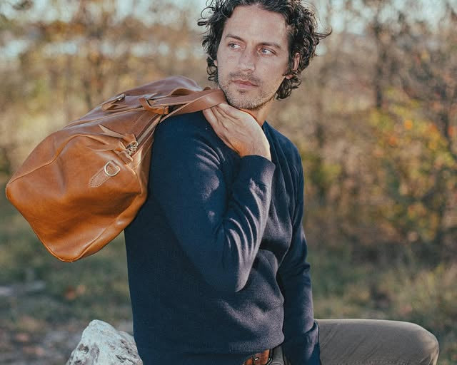 A man in a blue sweater holding a WP Standard’s weekender bag posing in a rural setting. 