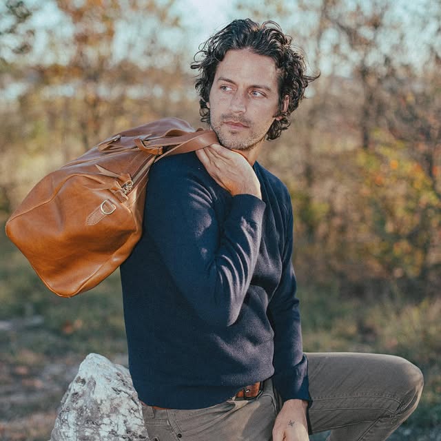 A man in a blue sweater holding a WP Standard’s weekender bag posing in a rural setting. 