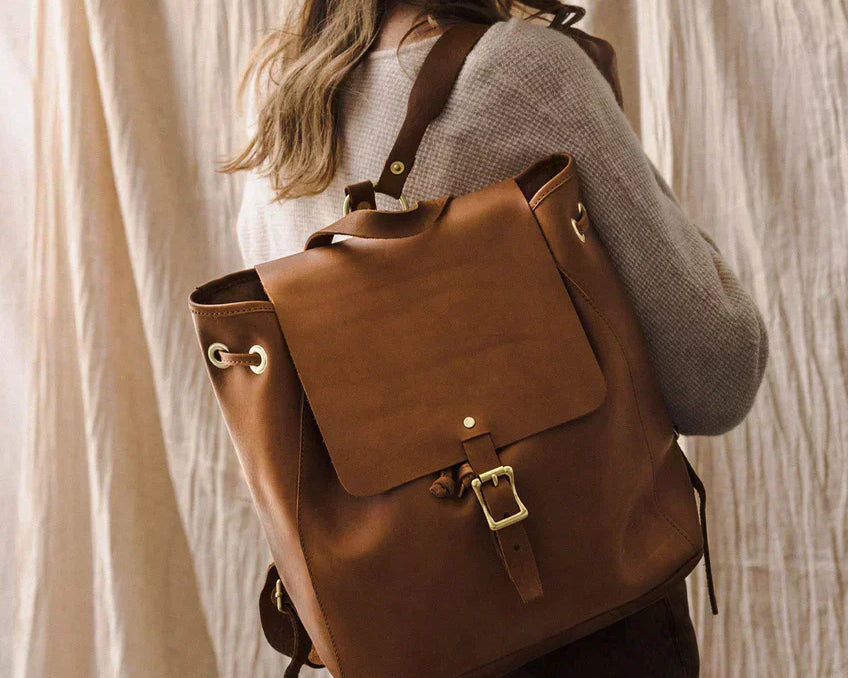 A woman wearing a brown leather backpack with a buckle closure and adjustable straps, paired with a wool hat and neutral outfit.