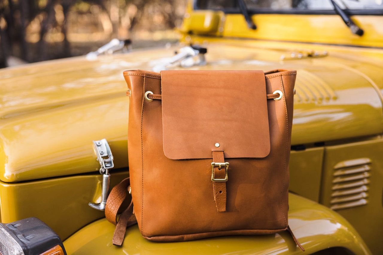 WP Standard tan leather backpack with a buckle closure, resting on the hood of a yellow vintage car.