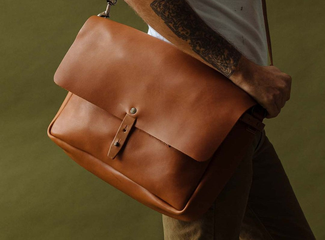 A man wearing a white t-shirt and khaki pants carries a tan leather Whipping Post messenger bag from WP Standard, shown against an olive green background.