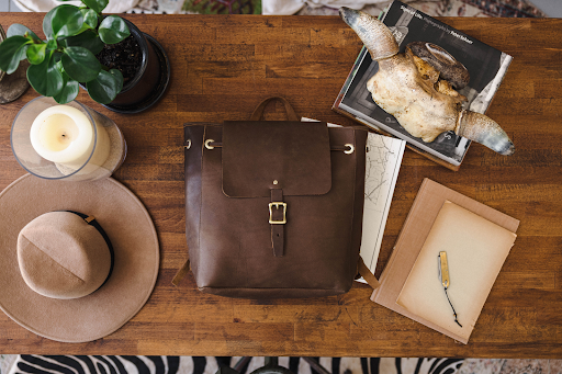 Top view of a brown leather backpack on a rustic wooden table surrounded by books, a candle, and decor items.