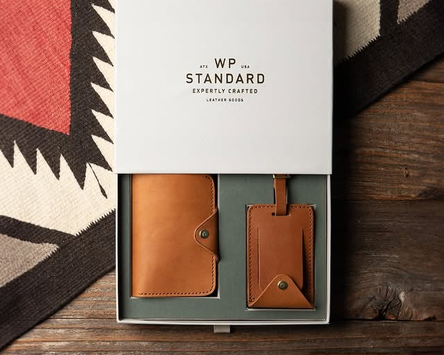 Tan leather wallet and matching luggage tag set from WP Standard displayed in a gift box on rustic wood.