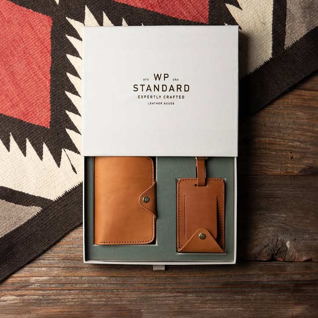 Tan leather wallet and matching luggage tag set from WP Standard displayed in a gift box on rustic wood.