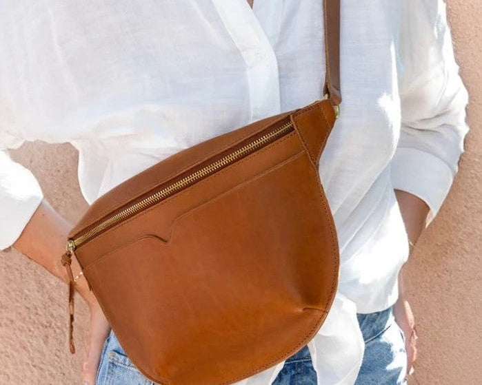 Brown leather crossbody bag worn by a person with a white blouse and denim jeans.