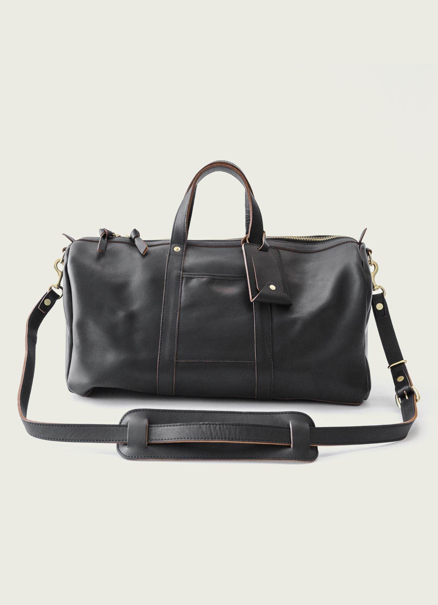 Elegant black leather duffle bag with gold-tone hardware, adjustable shoulder strap, and matching luggage tag – perfect for luxury travel and weekend getaways.